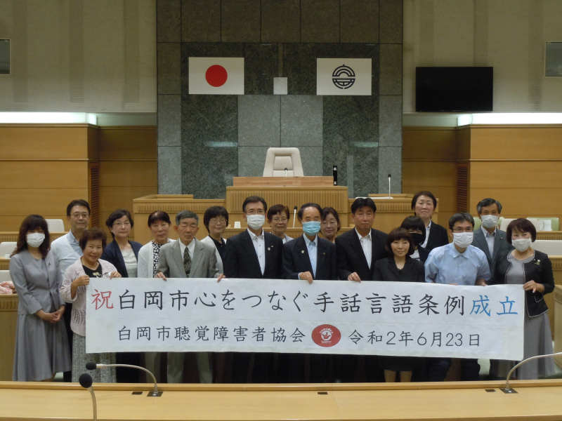 「祝白岡市心をつなぐ手話言語条例成立」と書かれた横断幕を手話言語条例制定検討会の参加者が持っている集合写真