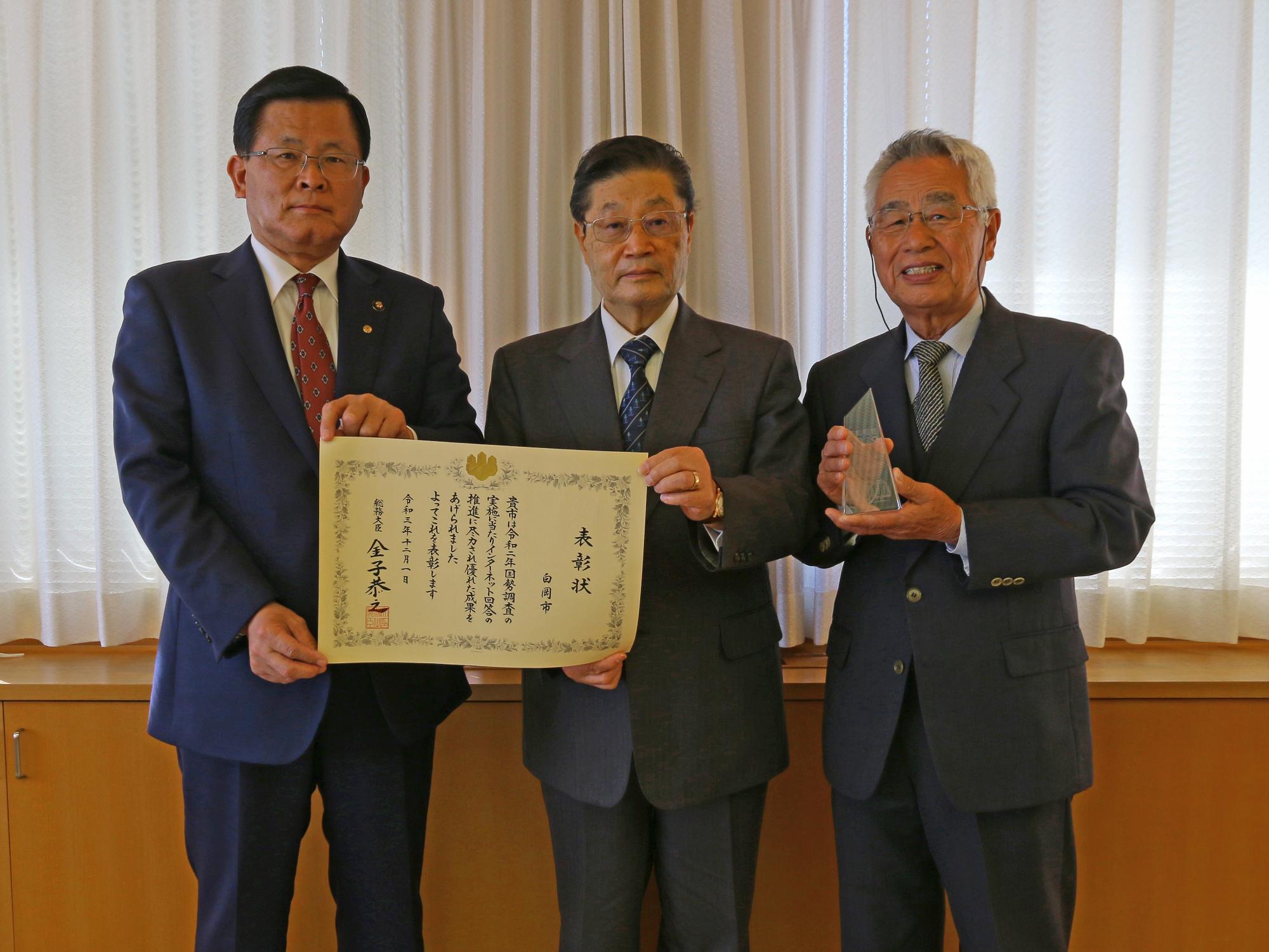総務大臣からの表彰状を持っている二人の男性と小さいトロフィーを持っている男性が笑顔で写っている記念写真