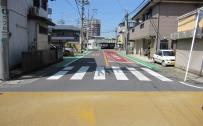 交差点の中心から見て、横断歩道の手前の道路が黄色く、奥から続く道路の一時停止線までが赤く、その道路の両端が歩道として緑色にペイントされている市道の写真