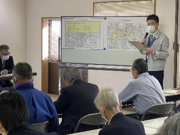 ホワイトボードに貼ってある資料を指しながら、マイクとバインダーを手に持って説明している1人の担当者の話を、説明会参加者たちが紙に書き込んでいる写真