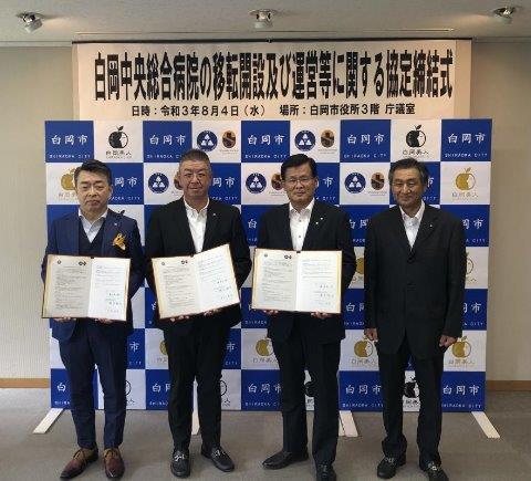 スーツを着て会見用のバックパネルの前に立ち協定書をもつ橋本委員長、中村会長、藤井市長と、江原議長の写真