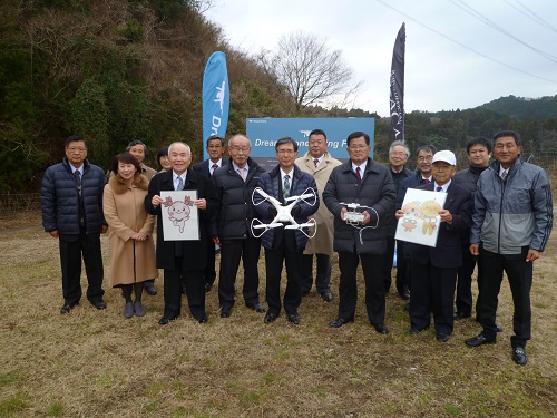自然を背景にしてドローンやイラストを持っている男性達と大勢の人達が写っている集合写真