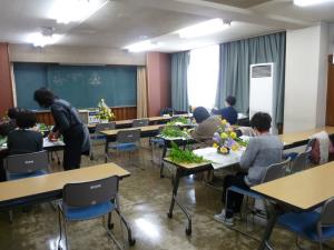 それぞれ席に座りフラワーアレンジメントの作品を作る受講者の方々と、1人の方の席に近づき直接アドバイスをする講師の方を部屋の後方から写した写真