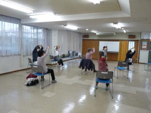 間隔をあけて椅子に座り両手を上にあげて輪を作り上半身だけ右にひねる運動をしている様子を後方から写した写真