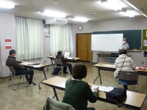 ホワイトボードの横に立ち話をする講師の方とテキストなどを見ながら話を聞く受講者の方々を部屋の後方から写した写真