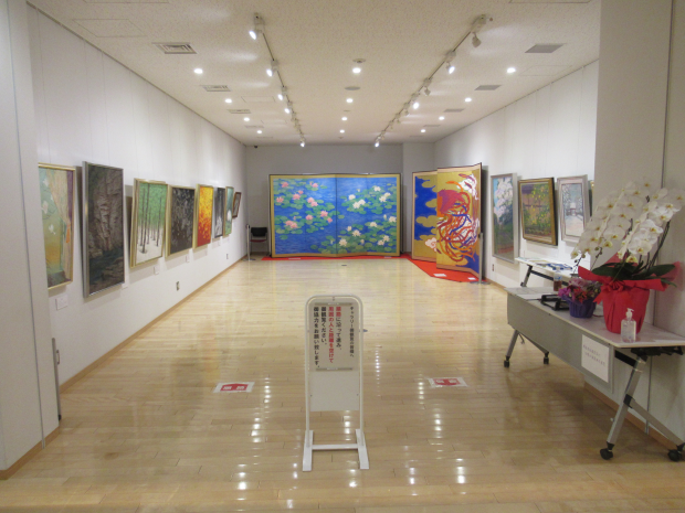 壁一面に絵や大きな屏風などが展示され、展示スペース手前の机に胡蝶蘭が置かれているギャラリー(企画展示室)の写真