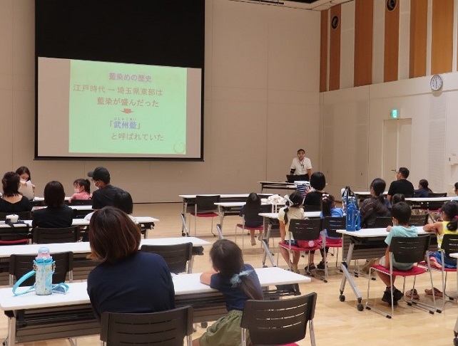 スクリーンに資料を映し説明する講師の方と書く自席につき説明を聞いている参加者の親子を後方から写した写真