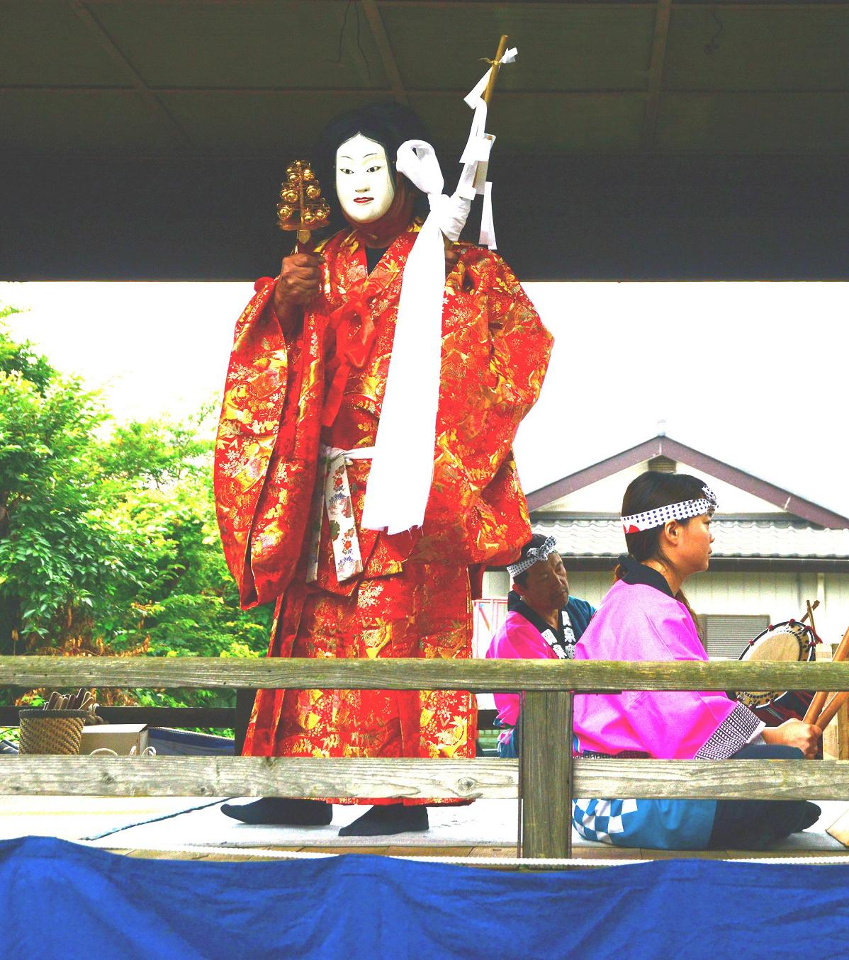 舞台でお面をつけ橙色の着物を着た巫女の姿をした踊り手が太鼓に合わせて舞っている岡泉鷲神社の神楽の写真