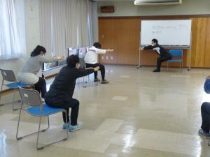 椅子に浅く腰掛け、両手をそろえて体ごと前に伸ばす運動をしている様子を後方から写した写真