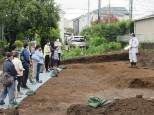 発掘調査現場で、右側に白い作業着を着た方が立ち、左側の地面にシートが敷かれた上に参加者たちが立って、作業着を着た方の説明を聞いている様子の写真