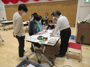 会場前方の机の周りに集まり学芸員の方の説明を聞く保護者の方と子供たちの写真