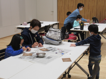 新聞紙を敷いた机の上で筆を使って色を塗っている親子の写真