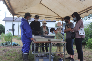 テントの下に設置された長机の上に並べられている出土遺物を覗き込んでいる参加者の男の子、男性、女性の写真