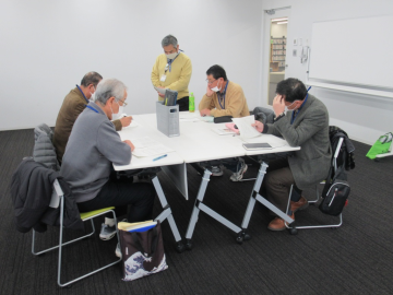 4人が席につき、1人の男性が席の近くに立って話し合いをしている様子を部屋の奥から撮影した写真