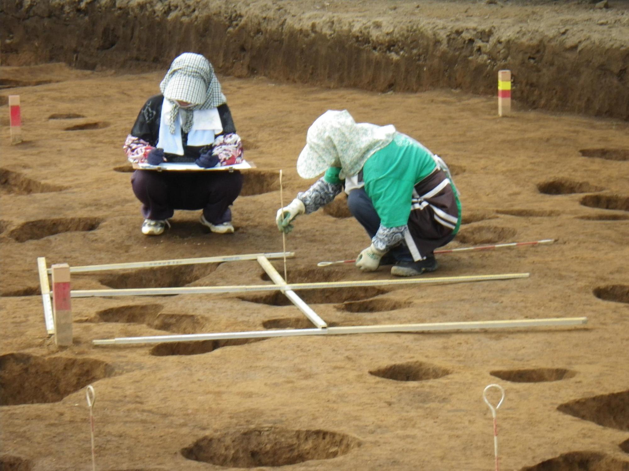 1名が木製の道具を使って穴の深さを計り、もう1名が記録を行っている作業員の写真
