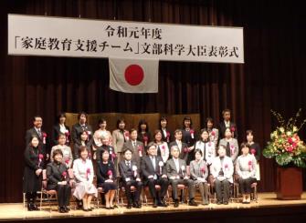 令和元年度「家庭教育支援チーム」文部科学大臣表彰式で檀上で並び記念撮影を行っている「町ぐるみん白岡」の関係者たちの写真