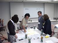 5名の参加者がテーブルの周りに集まり、、そのうち1名の男性がテーブルの上に置かれた模造紙を指さし、他の参加者たちが指さしている先を見ている写真