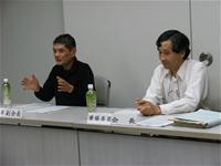 2名の男性が席に座っており、左側の男性が手ぶりを交えて話をしている様子の写真