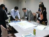 5名の参加者がテーブルの周りに立って集まり、模造紙をのぞき込むように話し合いが行われている様子の写真