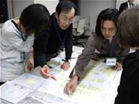 4人の参加者がテーブルの周りに集まり、そのうち2名の男性が模造紙を指さしたり、赤ペンで模造紙を指し話し合いが行われている様子の写真
