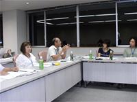 席に座っている参加者のうちの1人の男性が話している様子を、周りの参加者たちが話している男性の方を見て話を聞いている会議の様子写真