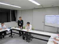 コの字型に並べられた席の前方側に男性が1名立ち、会議が行われている様子の写真