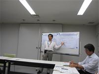 室内前方に置かれ、資料が貼られているホワイトボードの前に男性が立ち、ホワイトボードを指さしながら話をしている様子の写真