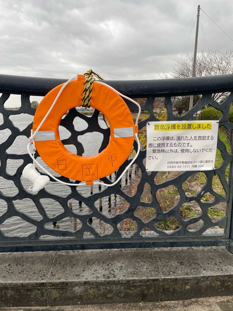 白岡市と書かれた救命浮環と、「救命浮環を設置しました。この浮環は、溺れた人を救助する際に使用するものです。緊急時以外は、使用しないでください。 白岡市都市整備部街づくり課公園担当 0480-92-1111 内線 204」と書かれた張り紙が柵に貼られている写真