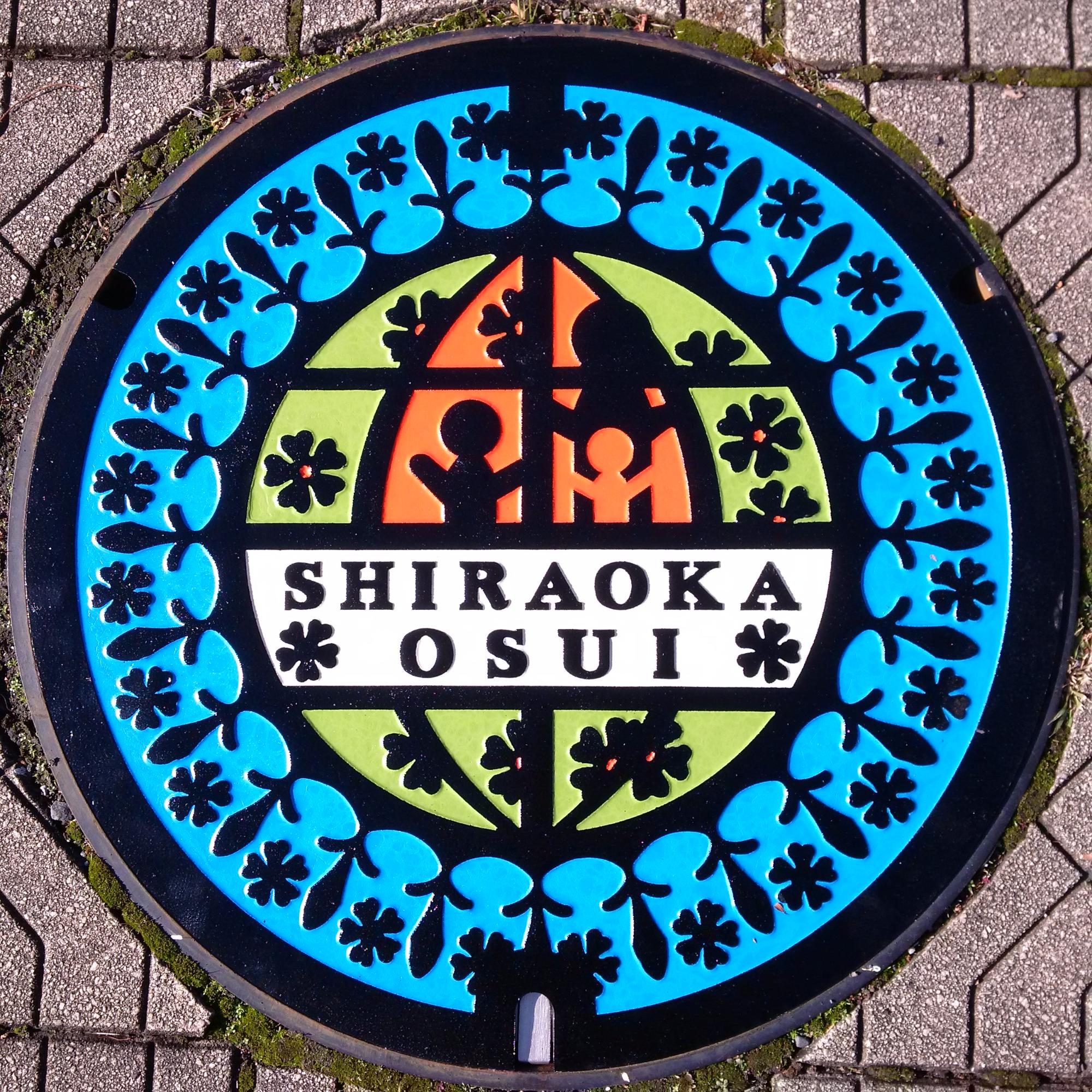 円の外側は青、中央に白地で「SHIRAOKA OSUI」と書かれその周りが緑とオレンジで着色されたカラーマンホールの写真