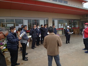 建物の前に集まって大勢の人達が紙に目を通して話を聞いている写真