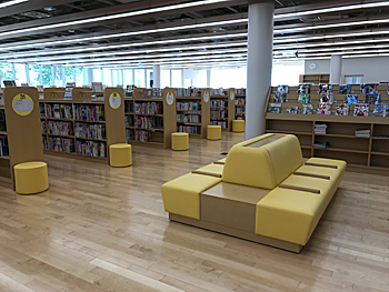 中央に背もたれのついた、背中合わせで8人座れる黄色のソファーが置かれており、奥には本棚がずらりと並んでいる、1階開架スペースの写真