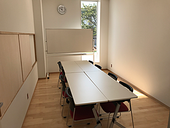 部屋の中央に8人掛けの机、奥にじゃホワイトボードが置かれており、部屋の奥の壁の右側の大きな窓から陽が差し込むグループ学習室の写真