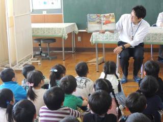 教室で職員の方が本を広げて紹介をし、子供たちが座って聞いている様子を後方から写した写真