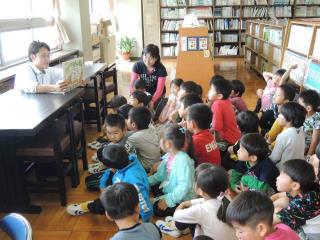 図書室で机の上に本を乗せて紹介する職員の方と、座って聞いている子どもたちの様子を横から撮影した写真