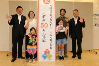 山下さんご家族と関係者2名の計6名が入館者50万人記念の立て看板をはさんで笑顔で手を振っている記念写真
