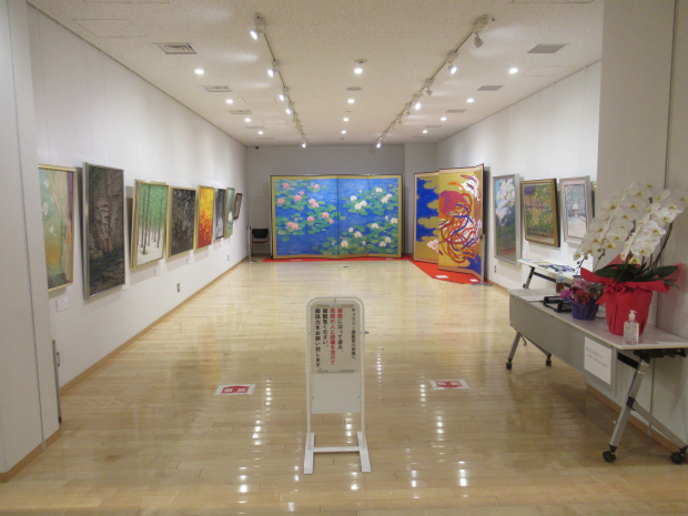 壁一面に絵や大きな屏風などが展示され、展示スペース手前の机に胡蝶蘭が置かれているギャラリー（企画展示室）の写真