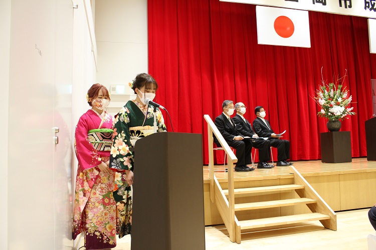 ステージ横の司会者席に立つ振袖を着た2人の新成人の写真