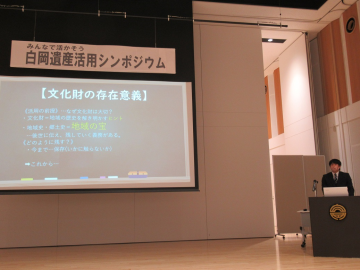 ステージ中央のスクリーンに資料を映し壇上から客席を見ながら発表をしている男性の写真