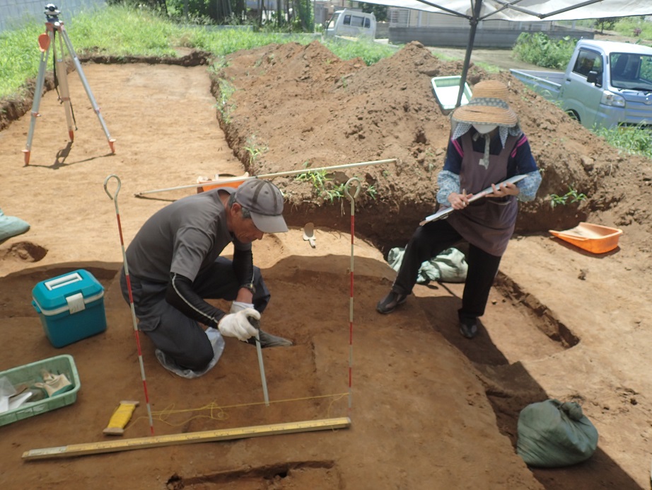 形どられた遺跡の跡に物差しなどを使い作業を行っている男性、図面のようなものを持っている女性、後方に光波測距儀が置いてある発掘調査の様子の写真