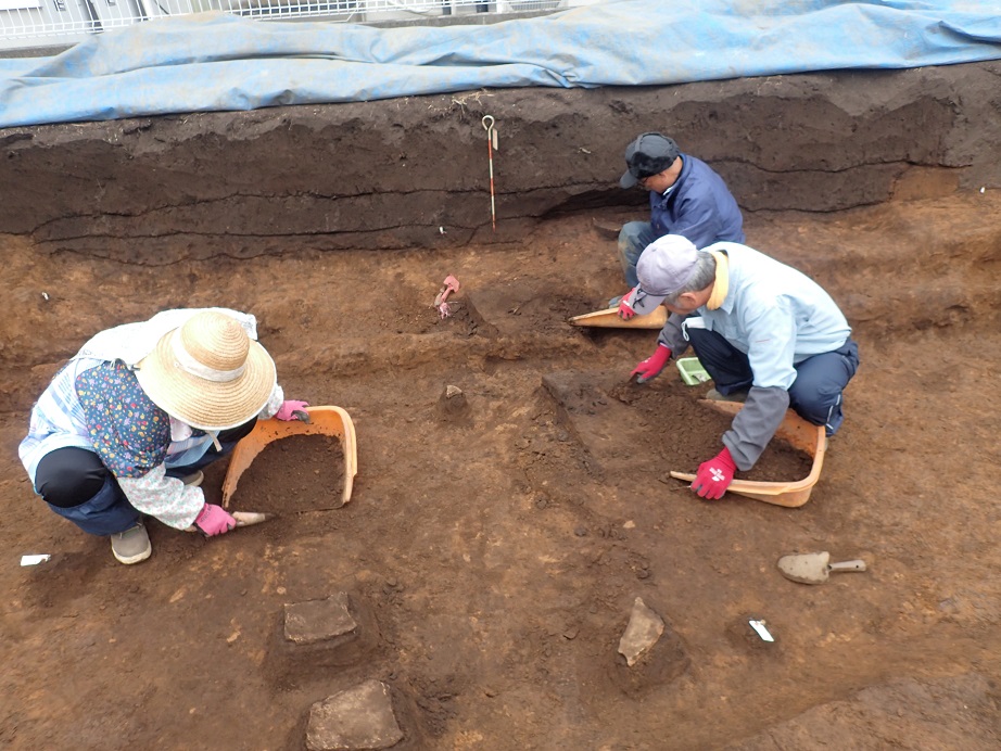 作業員3名が遺跡の中で、スコップで掘った土を手箕の中に入れながら、丁寧に発掘作業を行っている様子の写真