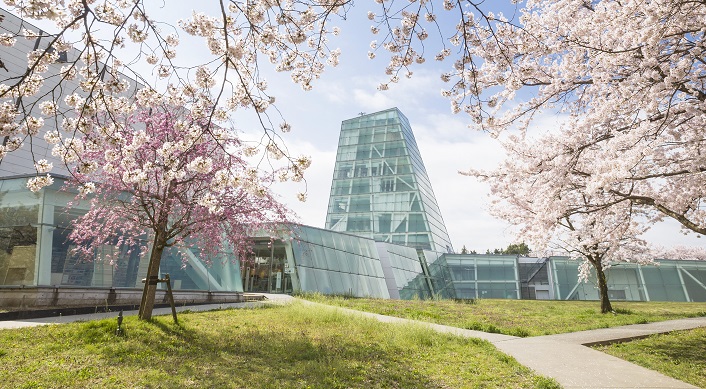 満開の桜の花が見える奥にある特徴的な形でガラスに覆われた日本工業大学LCセンターの外観写真