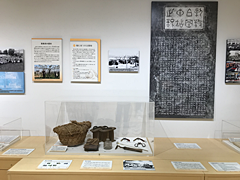 展示室の壁に資料や掛軸が展示され、壁の前の机にはにガラスケースの中に入った土器が展示してある写真