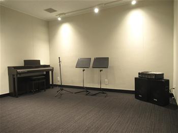 壁際に、ピアノ・マイクスタンド・譜面台2つ・スピーカー置かれている音楽スタジオの写真
