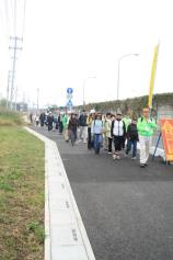 緑のジャンパーを着ている人を先頭に、たくさんの参加者の人達が並びながら道路の脇を歩いて避難誘導訓練を行っている様子の写真