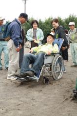 参加者の人達が、車椅子に乗り疑似体験をしている男性を囲んでいる写真