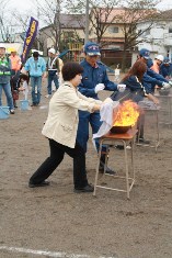 各机の前で炎が舞い上がった天ぷら鍋に女性が濡れたシーツをかぶせ、横に消防隊員の方が立ち指導している消火訓練の写真