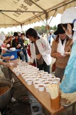 紙コップに入ったすき焼き丼と筑前煮丼が長机に並べられ、たくさんの人達が机の前に並んで見学をしている写真