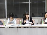3名の参加者が席に座っており、両側の参加者が資料に目を向け、中央の女性が話をしている様子の写真