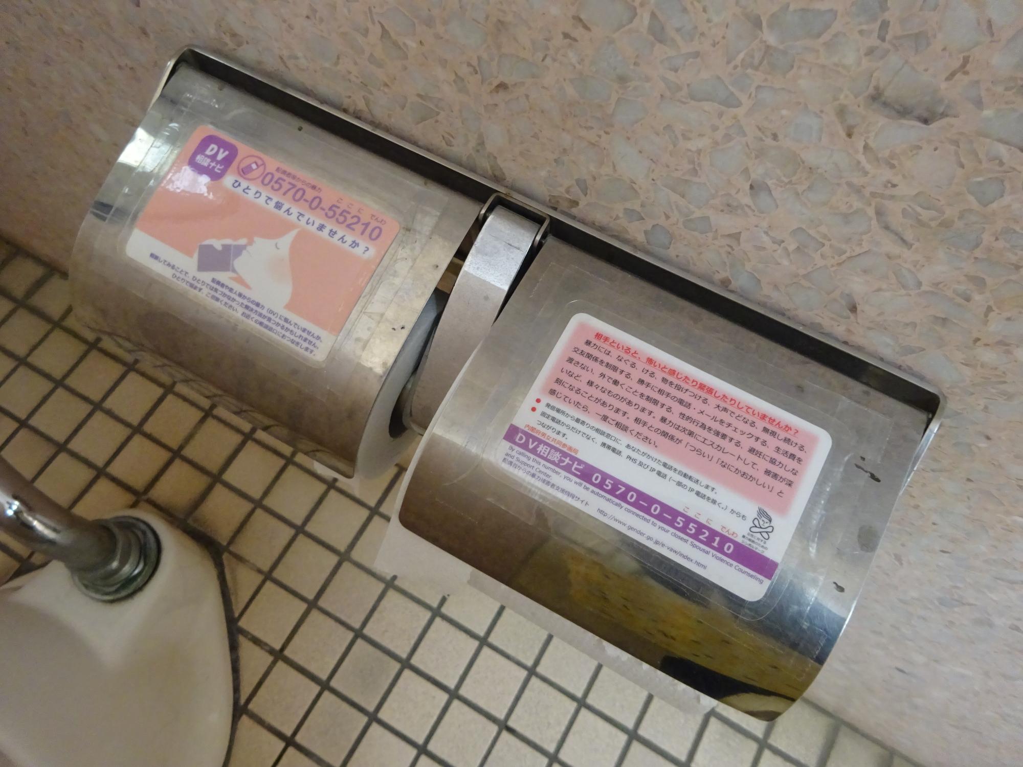 和式トイレ内のトイレットペーパーホルダーのシルバーのカバー部分にDV相談ナビカードが貼られている写真
