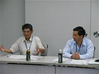2名の男性が席に座り、左側の男性が手ぶりを交えて話をしている様子を右側の男性が見ている写真
